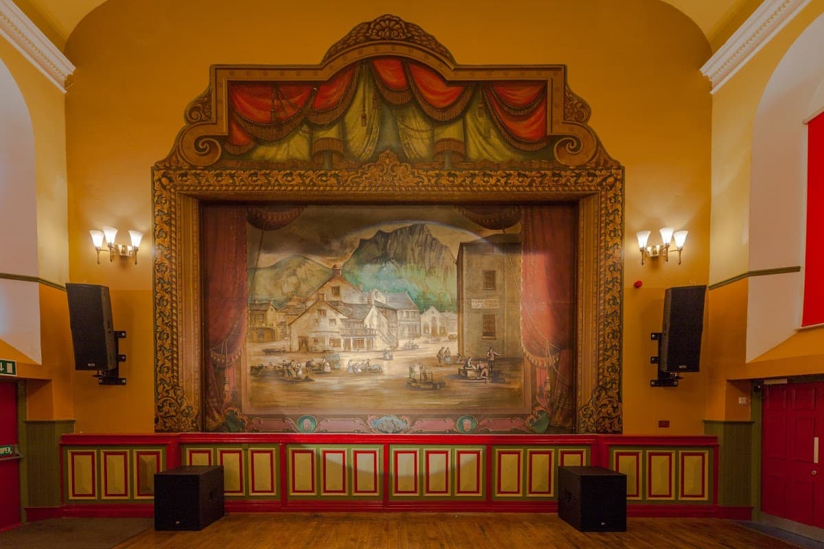 Theatrical stage with a large painted backdrop, ornate curtains, and wall sconces in a vibrant community hall.