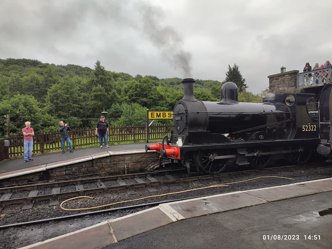 Steam locomotive at Embsay Railway Station with passengers observing and lush greenery in the background.