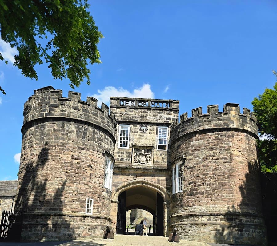 Historic Skipton Castle features stone walls, turrets, and a grand entrance under a bright blue sky.