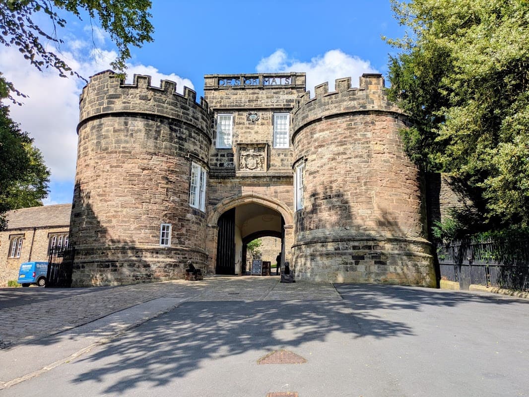 Historic stone entrance with towers, trees lining the path, and a blue sky in Skipton Castle Woods, Yorkshire.