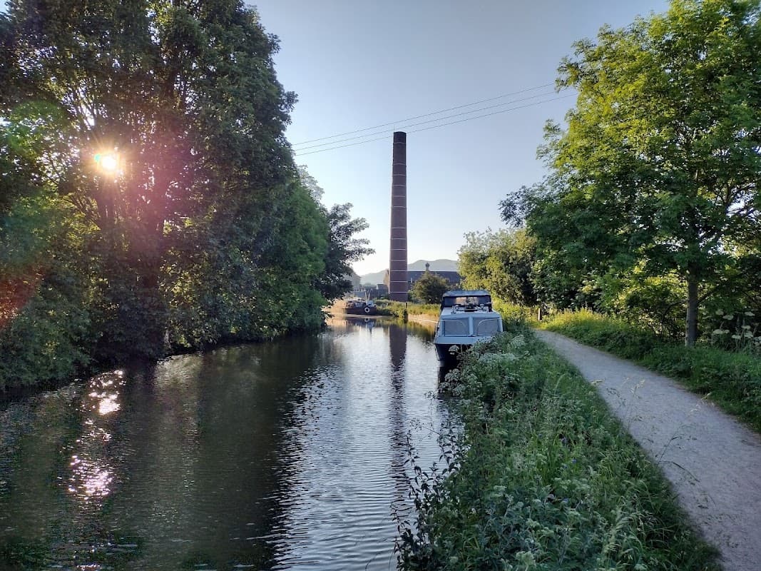 A serene canal scene in Skipton, Yorkshire, with a boat, lush greenery, and a tall brick chimney in the background.