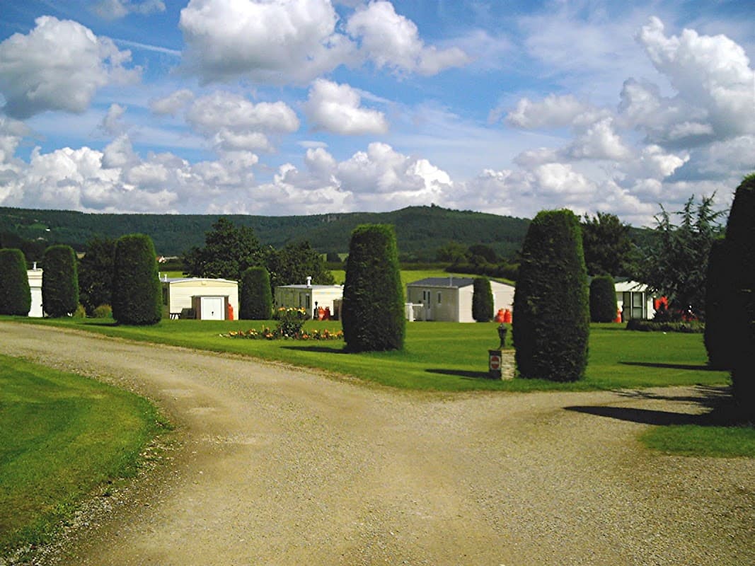 Gravel path leads through manicured lawns and tall hedges, with holiday cabins nestled against a backdrop of rolling hills.