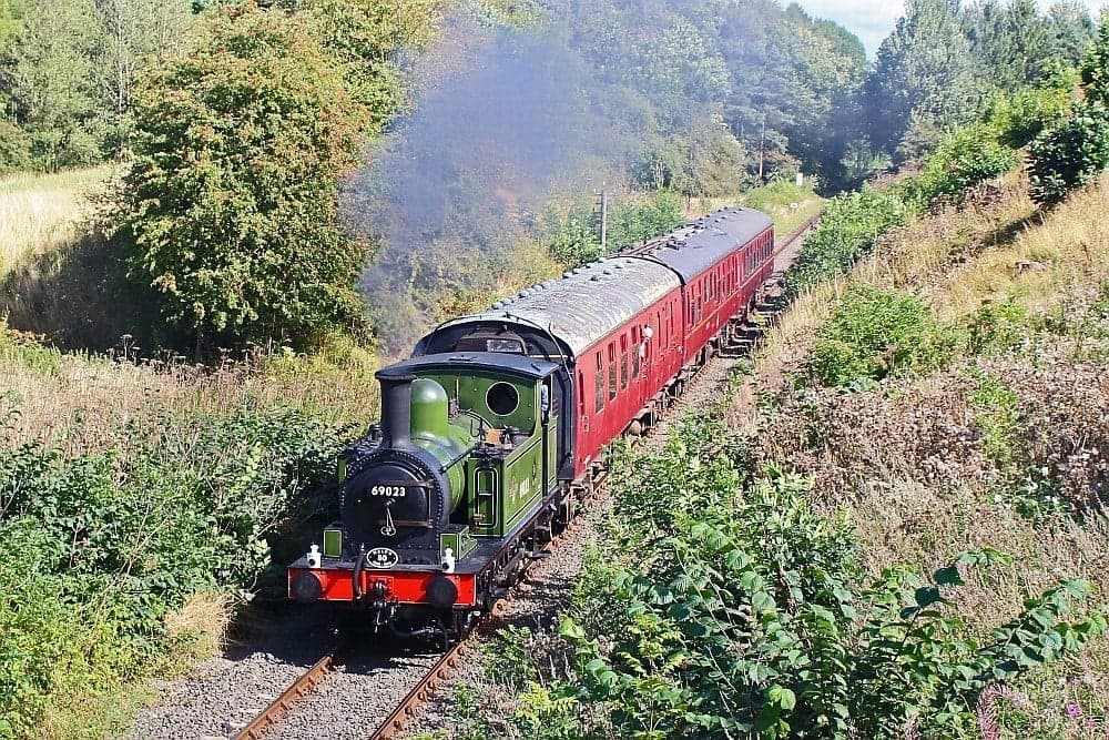 Historic steam train with green engine and red carriages travels through lush countryside surrounded by trees and shrubs.