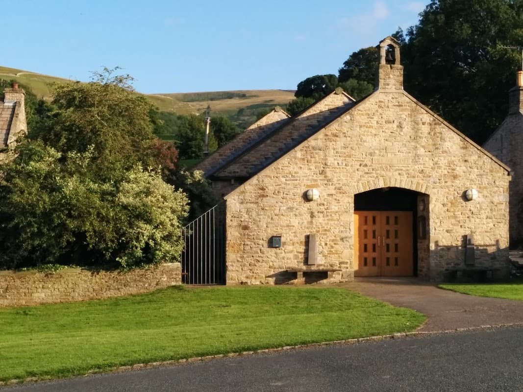 Stone building with a bell tower, green lawn, and trees, set against a backdrop of rolling hills in West Burton.