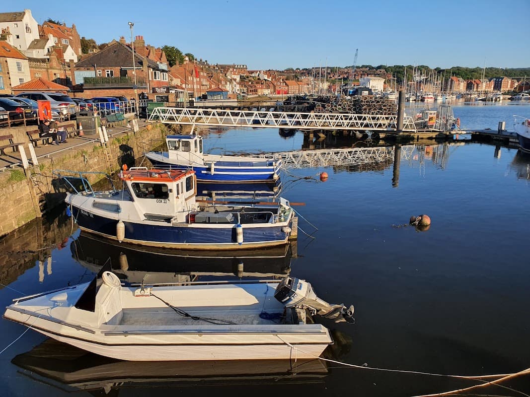 Boats moored at a dock in Whitby, Yorkshire, with buildings and hills in the background under a clear blue sky.