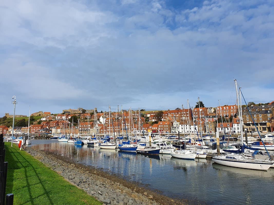 Harbor with moored sailboats, colorful buildings on the hillside, and a cloudy blue sky in Whitby, Yorkshire.