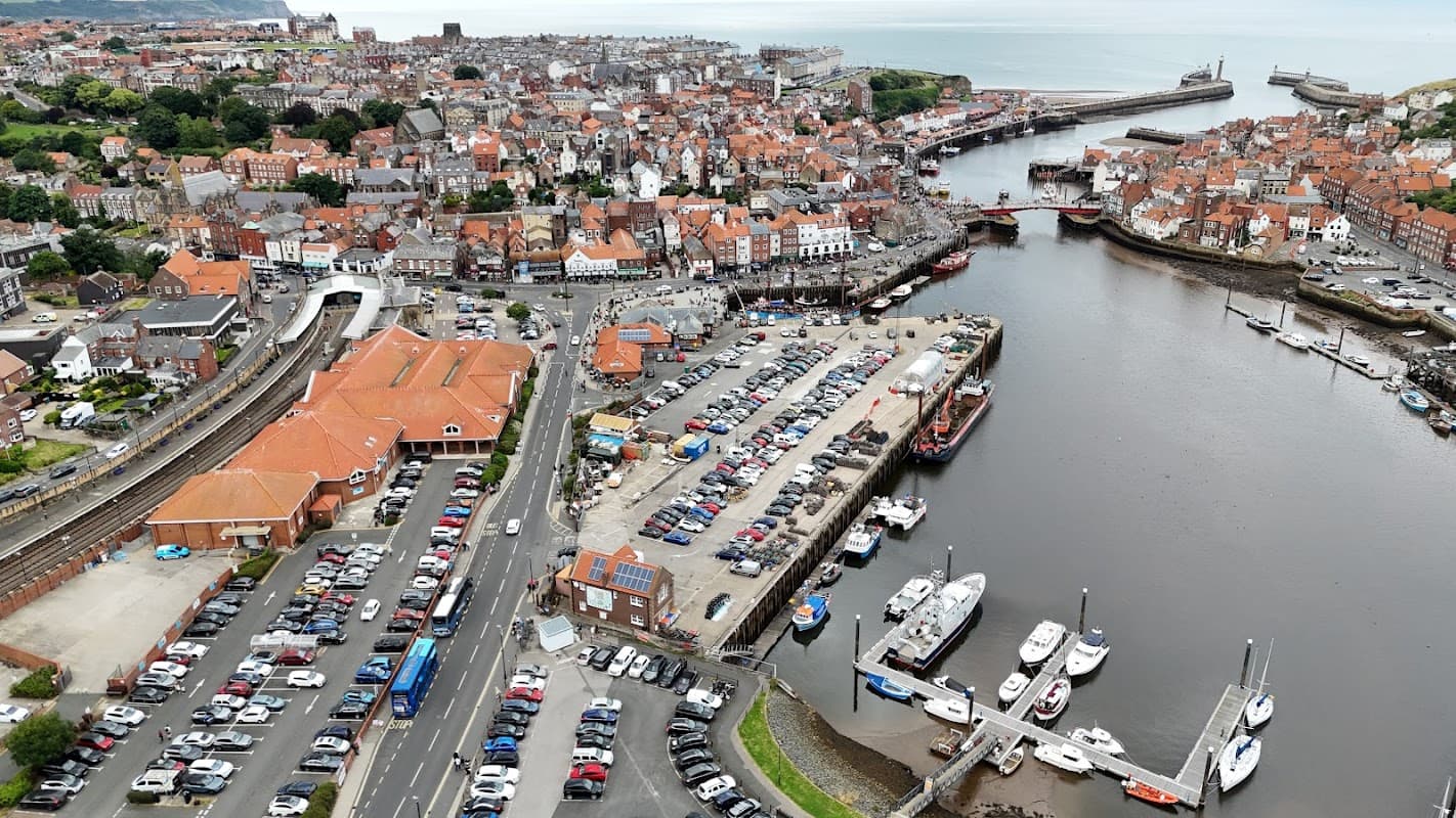 Aerial view of Marina Front Car Park in Whitby, featuring cars, boats, and the coastal town with red-roofed buildings.