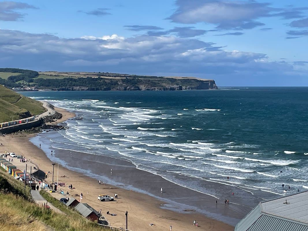 North Terrace Car Park overlooking a sandy beach with waves, colorful beach huts, and cliffs in the background.
