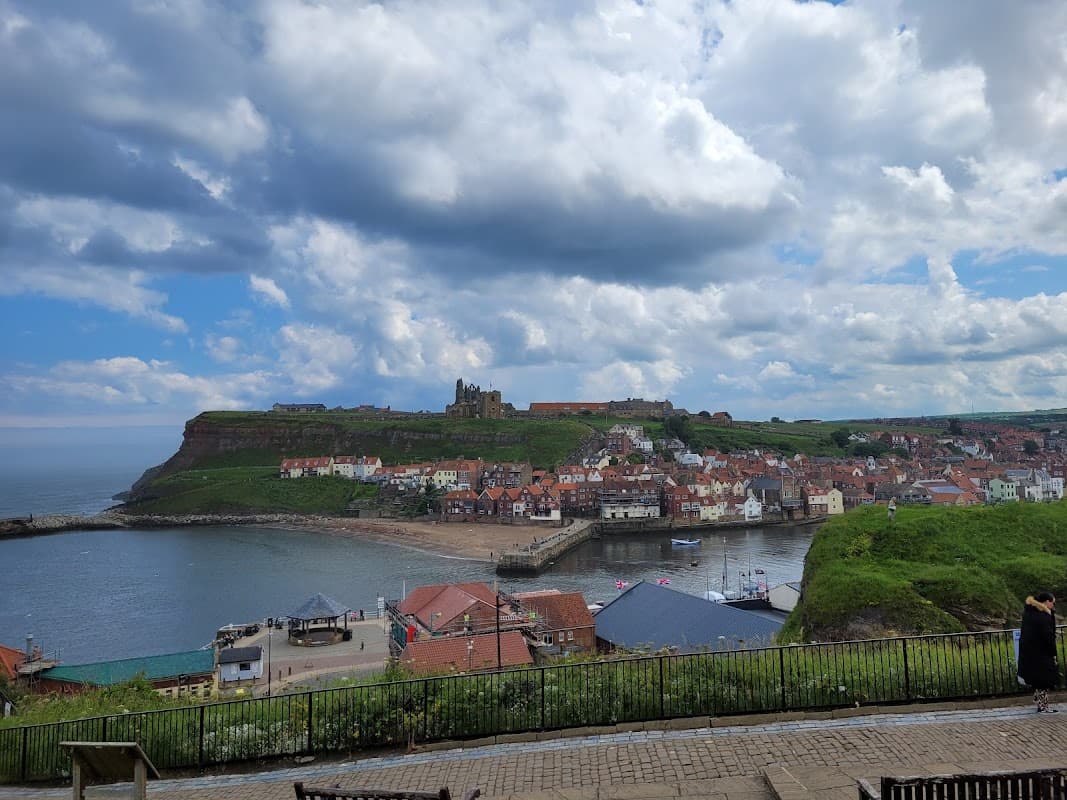 Panoramic view of Whitby with colorful buildings, a harbor, and cliffs under a partly cloudy sky.
