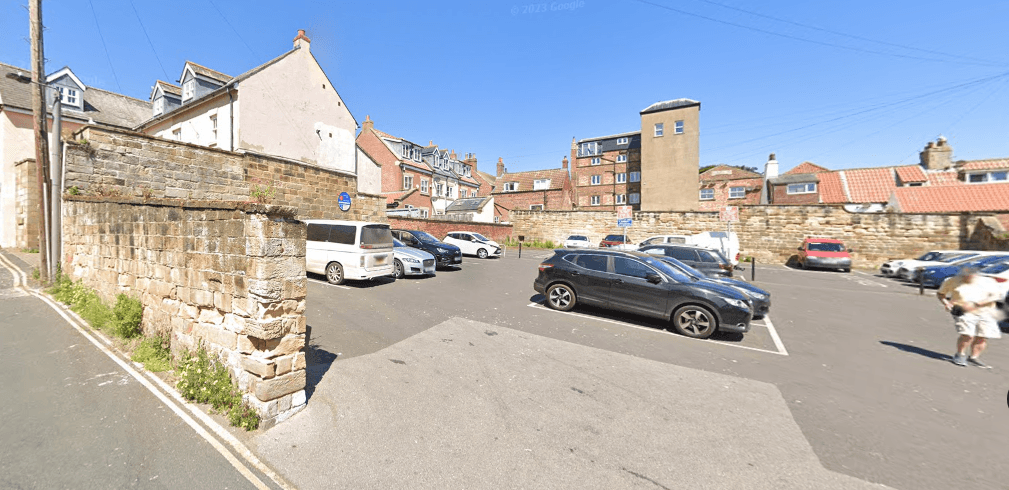 Car park with multiple vehicles, stone walls, and buildings under a clear blue sky in Whitby, Yorkshire.