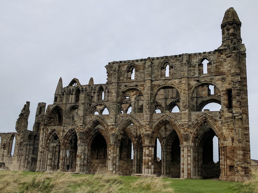Ruins of Whitby Abbey with stone arches and towers, set against a cloudy sky and grassy foreground.