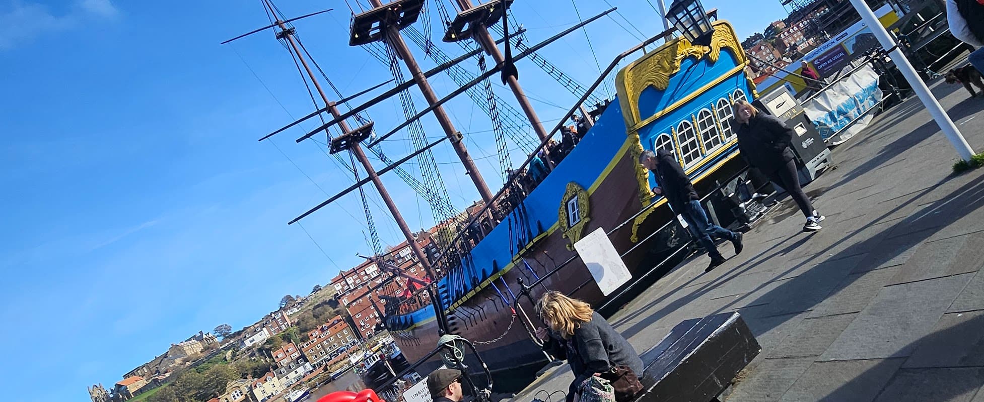 Colorful pirate ship docked at Whitby, with people walking along the waterfront and scenic hills in the background.