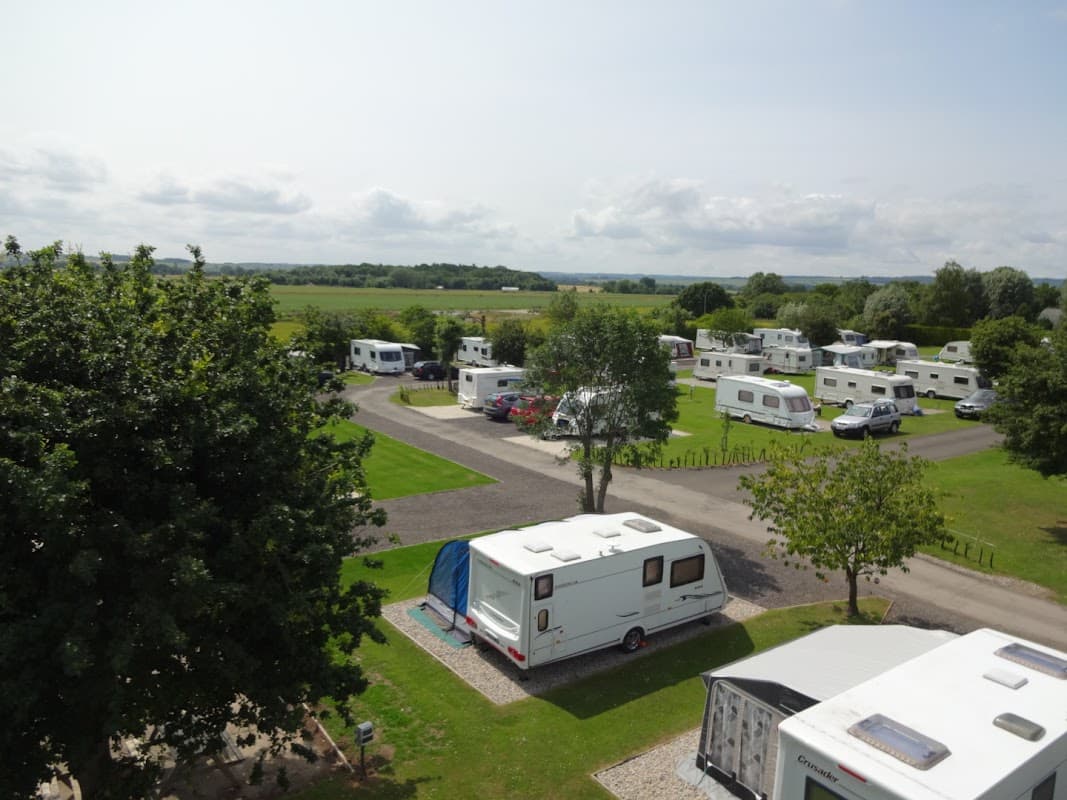 Caravans and motorhomes parked on a grassy campsite with trees and open fields in the background.