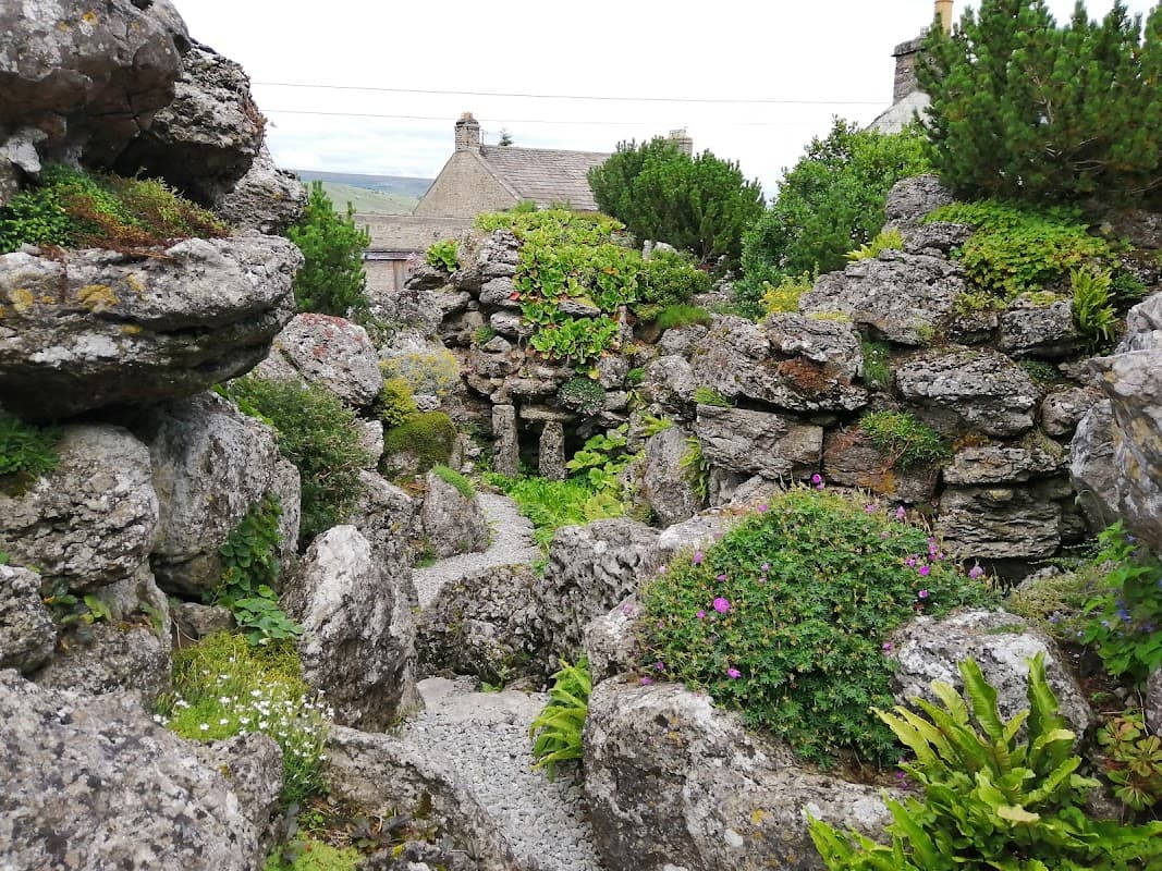 Lush greenery and stone formations create a serene pathway in the Aysgarth Edwardian Rock Garden, surrounded by buildings.