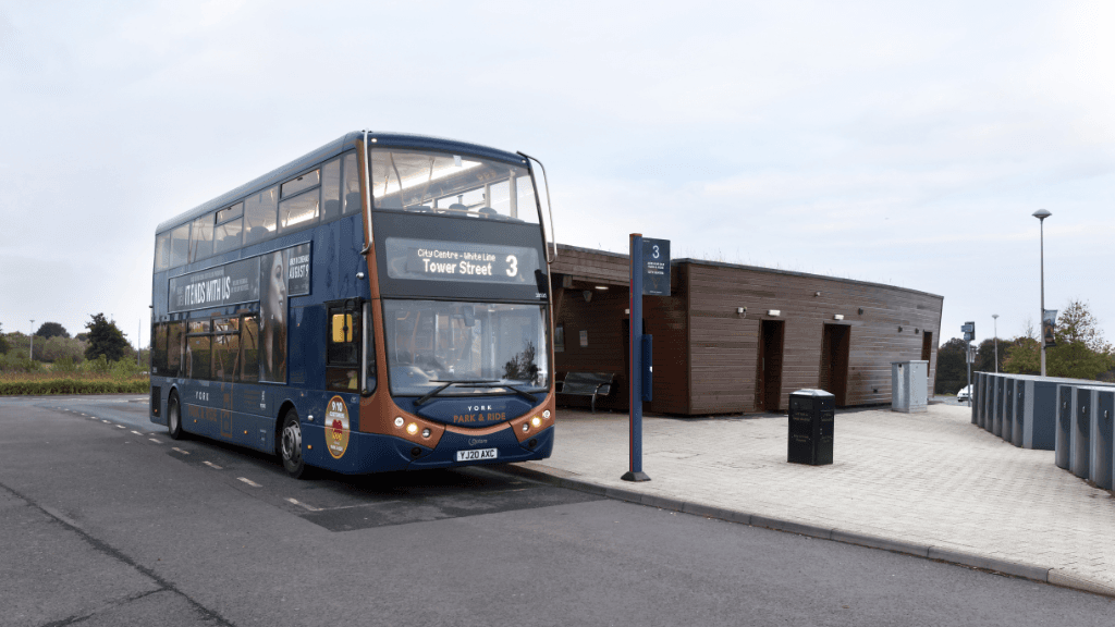 A double-decker bus at a park and ride stop, with a modern building and signage for route 3 to Tower Street.