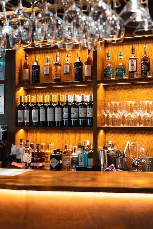 Wooden bar with shelves displaying various bottles of spirits and glasses, illuminated by warm lighting.