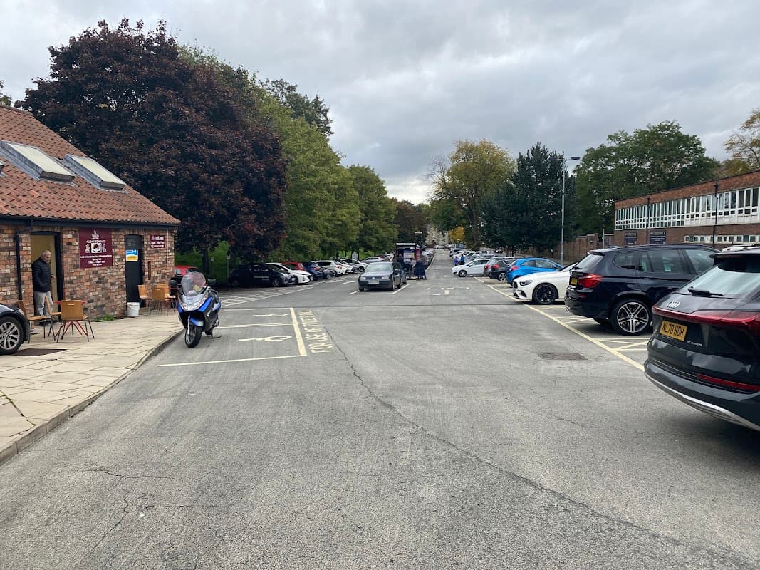 Pay & Display car park with parked cars, trees lining the sides, and a building with a red roof in York, Yorkshire.