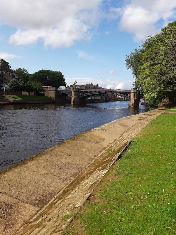 View of a riverbank in York with a bridge, greenery, and a cloudy blue sky above.