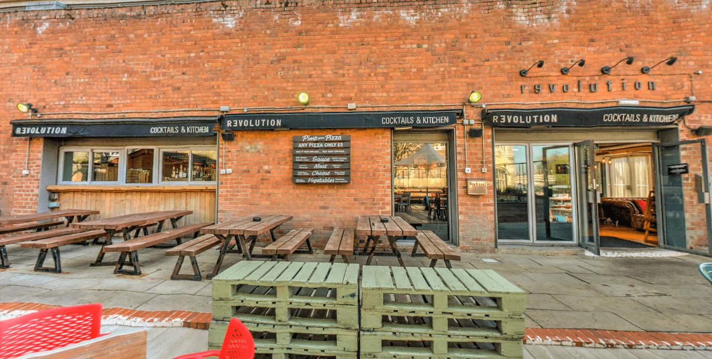 Outdoor seating area with wooden benches, a bar entrance, and a brick wall featuring the "Revolution" sign.