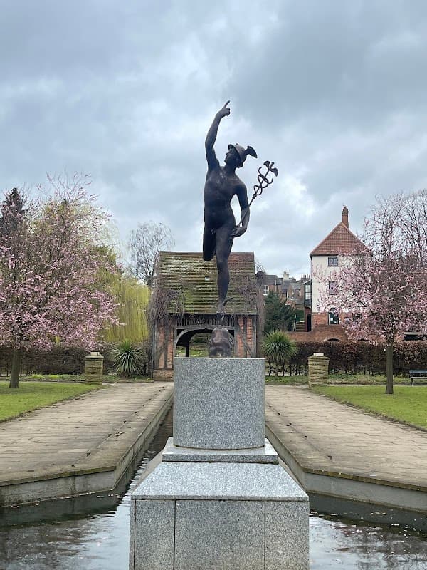 A bronze statue of a figure with a staff stands on a pedestal, surrounded by blooming trees and a pond.