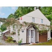 Cream-colored two-story garden cottage with ivy-covered trellis, brown garage door, and lush greenery surrounding.
